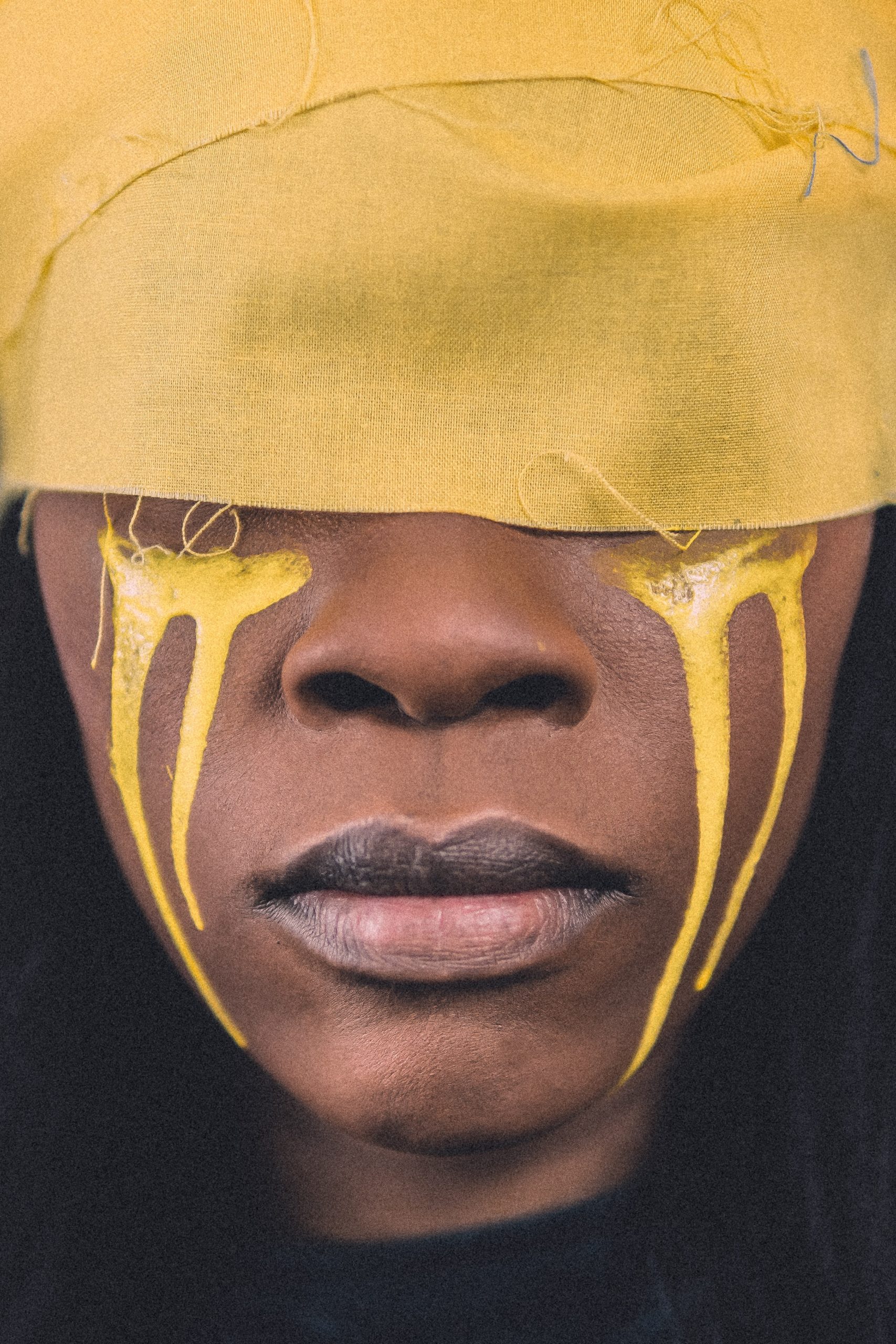 Black woman with a yellow strip around her eyes. Yellow paint like tears is painted down her face to her chin.